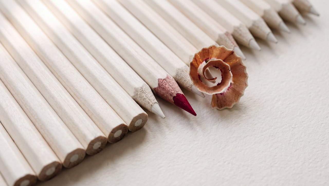 A row of white pencils, one sharpened to a red point beside its curled shavings